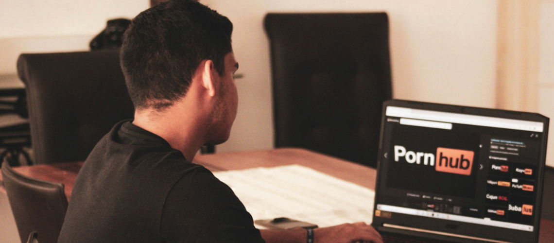 Man sitting at a table using a laptop with a pornography website open, highlighting the private nature of porn consumption and its connection to real-world exploitation.