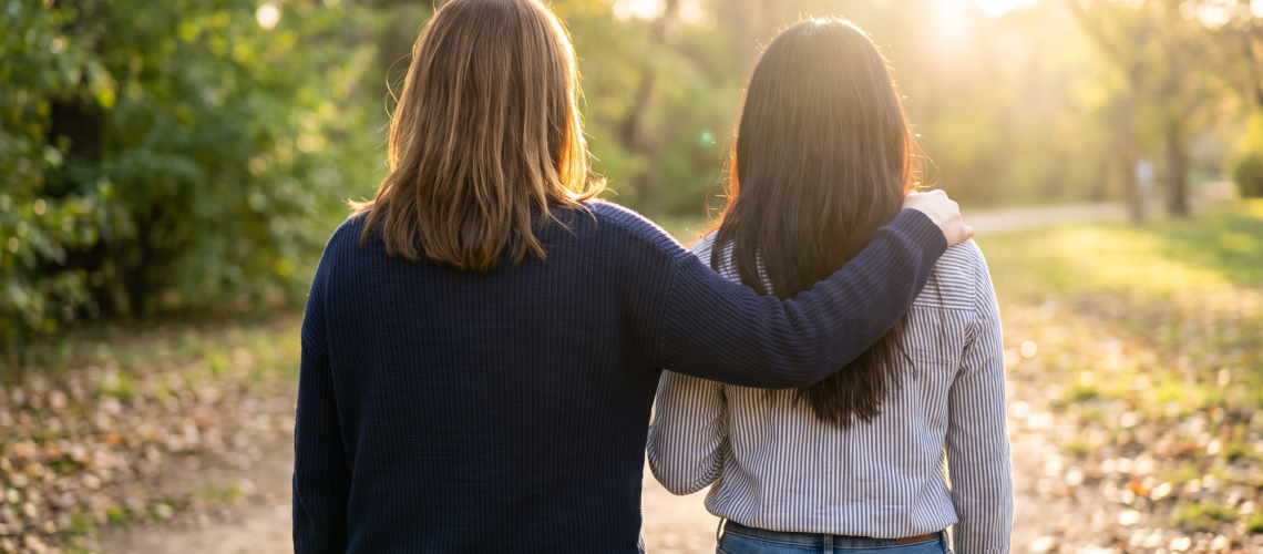 Two women seen from behind walking together on a sunlit park path, one with an arm around the other's shoulder, symbolizing mentorship and community support for survivors