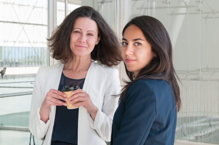 Content colleagues relaxing and looking at camera in office hall. Attractive women standing and drinking coffee. Break concept.